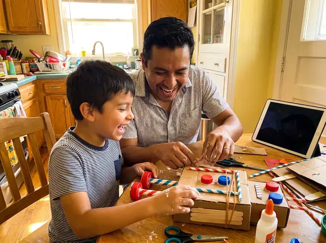 Papá e hijo riendo mientras arman un robot de cartón en la mesa de la cocina